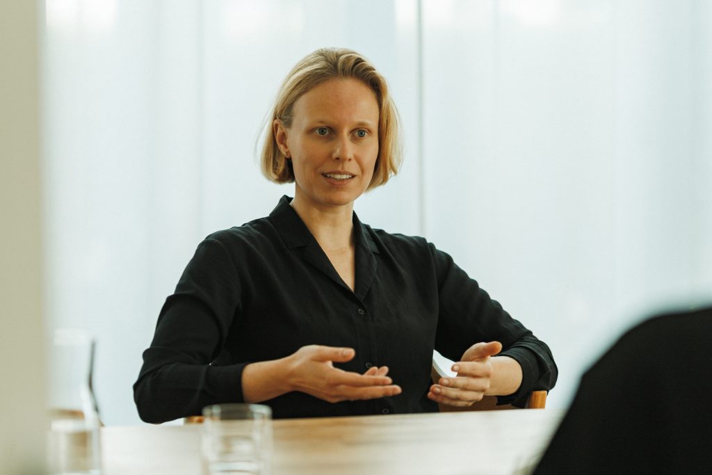 Kerstin Stock, eine blonde, leicht lächelnde und dunkel gekleidete Frau, sitzt an einem Holztisch und hält ihre beiden Hände in einer erläuternden Geste vor ihrem Oberkörper.