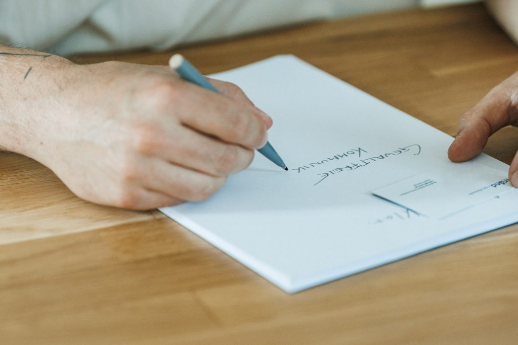 Eine Hand hält eine friånd Visitenkarte, während eine andere Hand das Wort "Gewaltfreie Kommunikation" auf ein weißes Blatt Papier in Großbuchstaben schreibt.