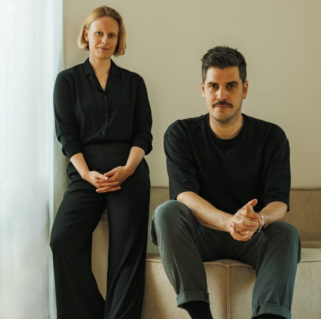 Portrait einer stehenden Frau, Kerstin Stock, und eines sitzenden Mannes, Tobias Schneider, beide schwarz gekleidet, mit neutralem bis freundlichem Gesichtsausdruck.