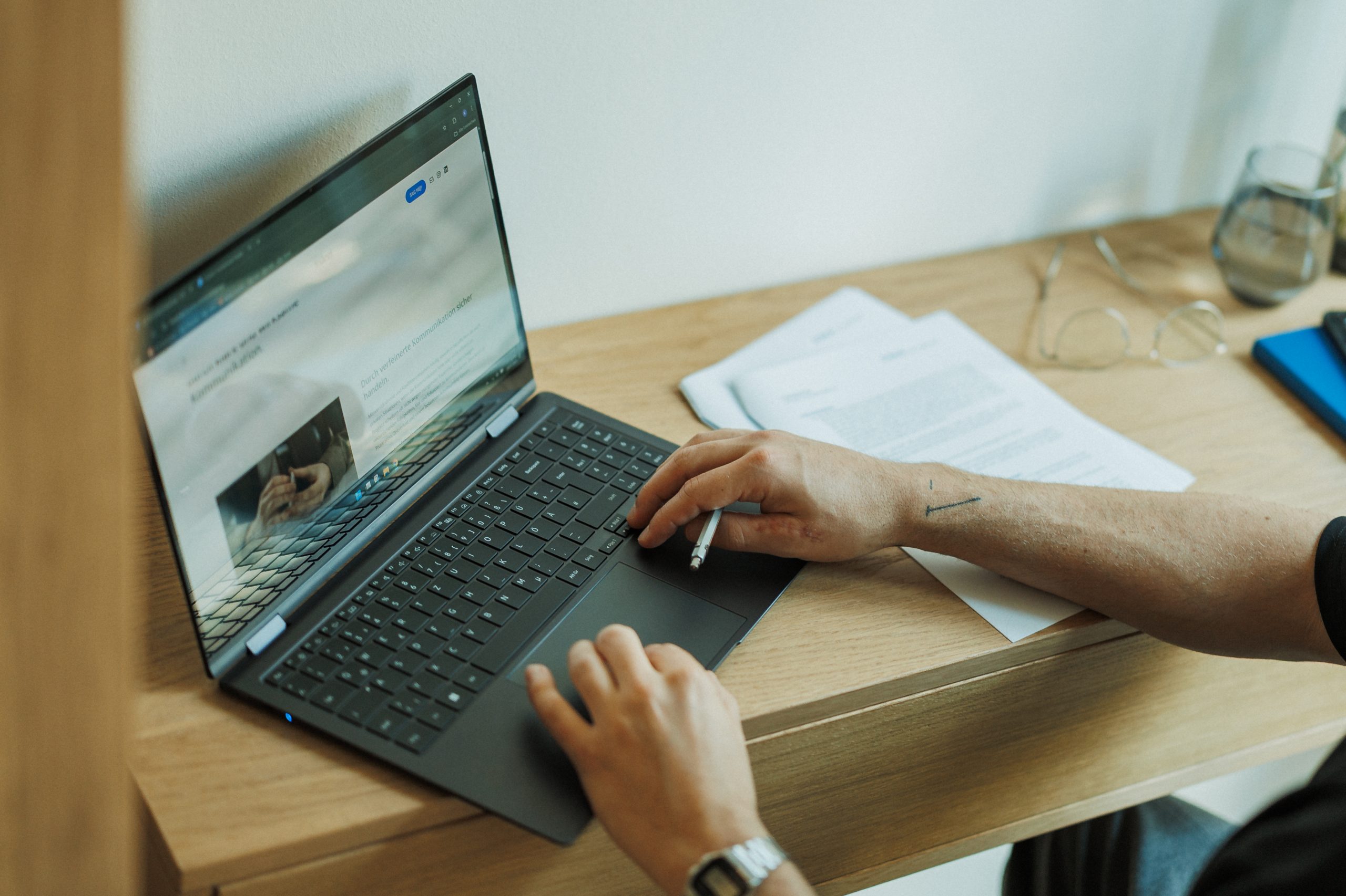 Ein schwarzer Laptop auf einem Holztisch, eine Brille, ein Glas, lose Papiere sowie ein paar Hände, die auf der Tastatur des Laptops abgelegt sind.