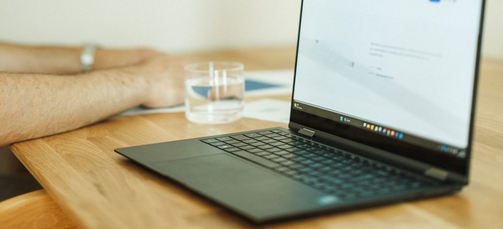 Ein eingeschalteter schwarzer Laptop auf einem Holztisch in einer Seminar-Situation. Daneben ein paar Hände und ein Wasserglas.