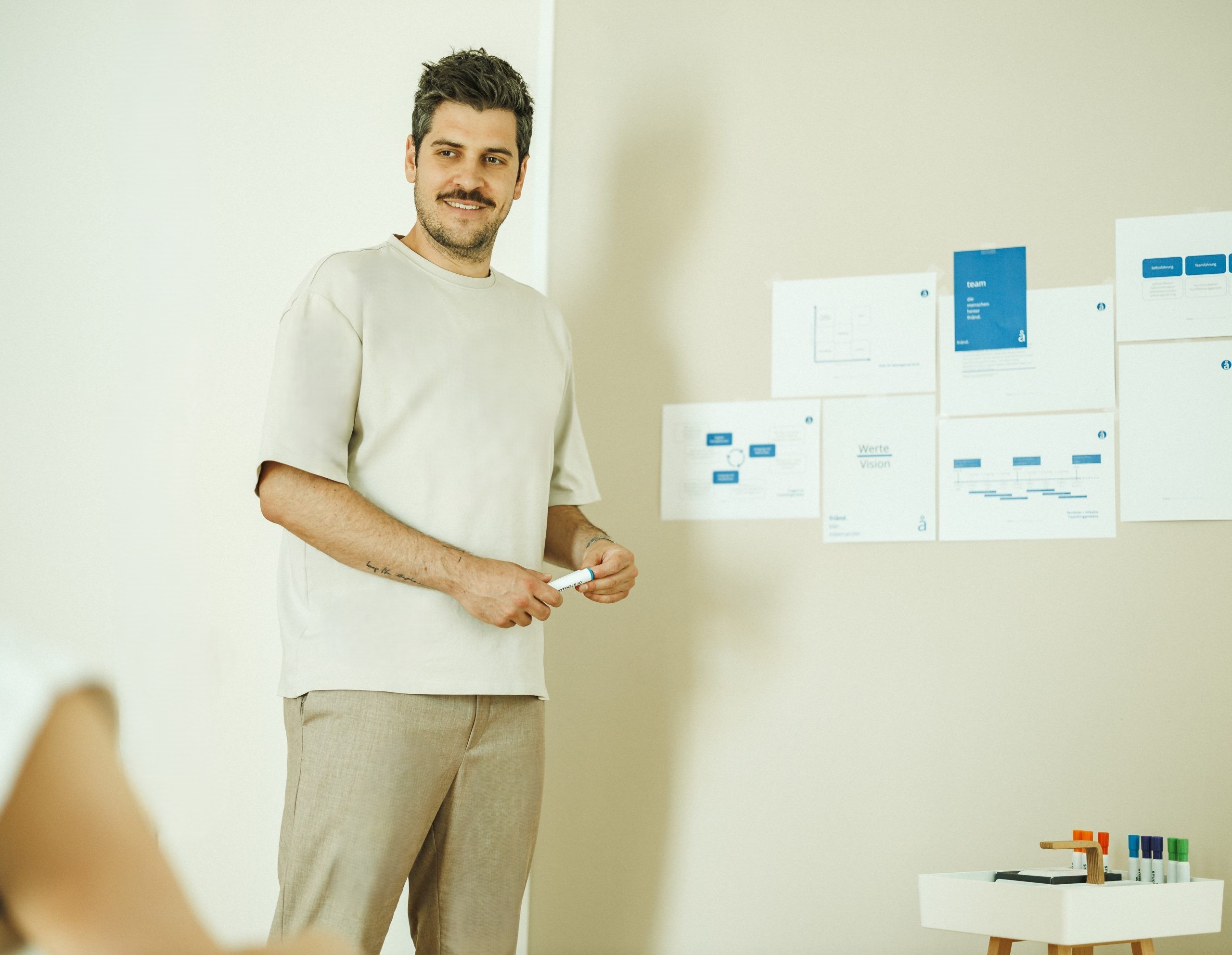 Tobias Schneider, ein lächelnder Mann, hält einen Stift in der Hand und steht vor einer Wand mit Postern, während er ein Seminar hält.