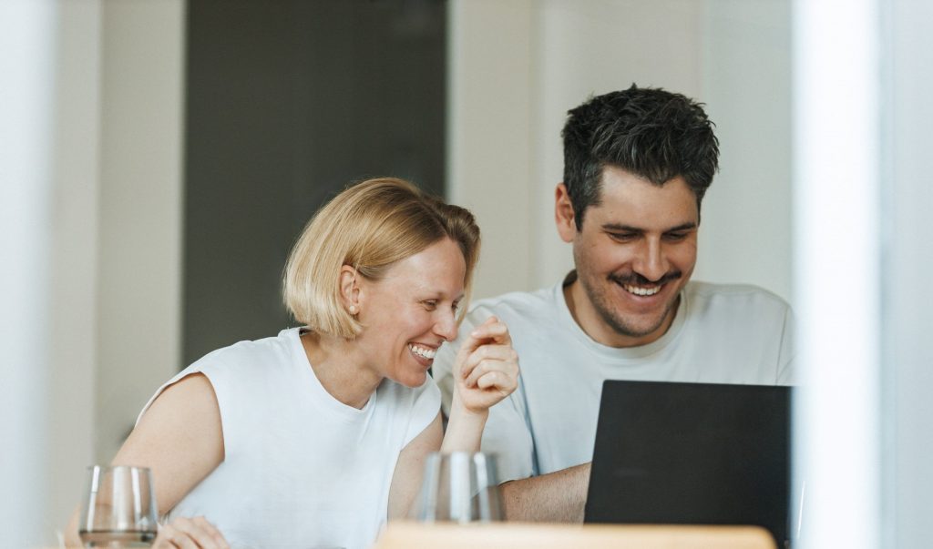 Eine blonde Frau und ein braunhaariger Mann schauen lachend auf einen Laptop-Bildschirm, während sie nebeneinander an einem Tisch sitzen.