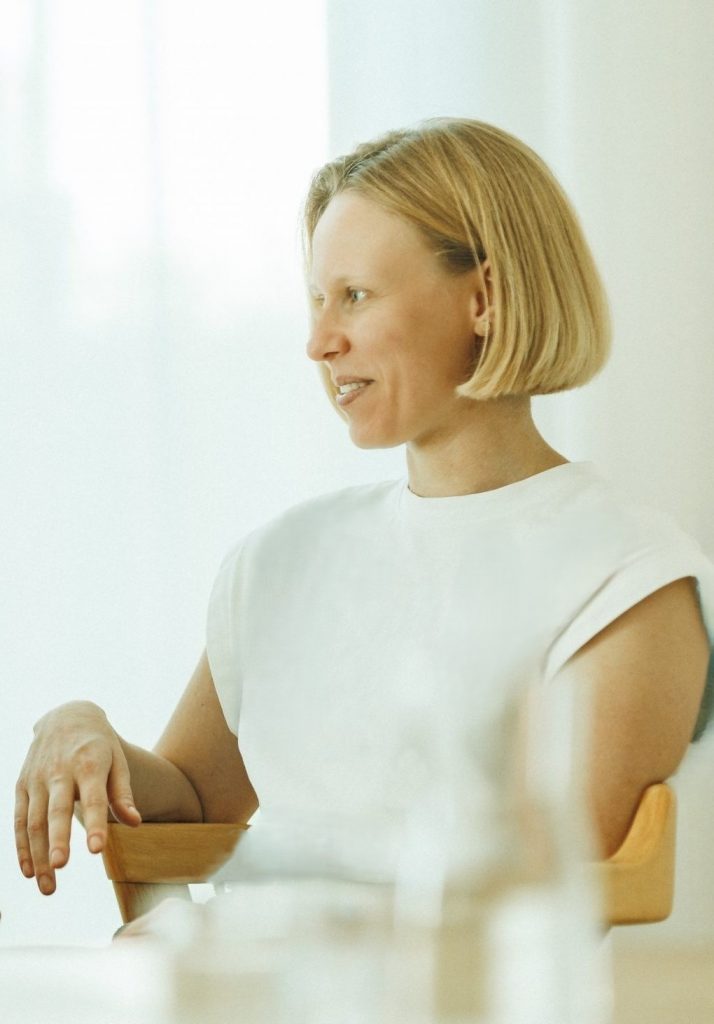 Kerstin Stock, eine blonde und hell gekleidete Frau, von der Seite gesehen, sitzt auf einem Stuhl.