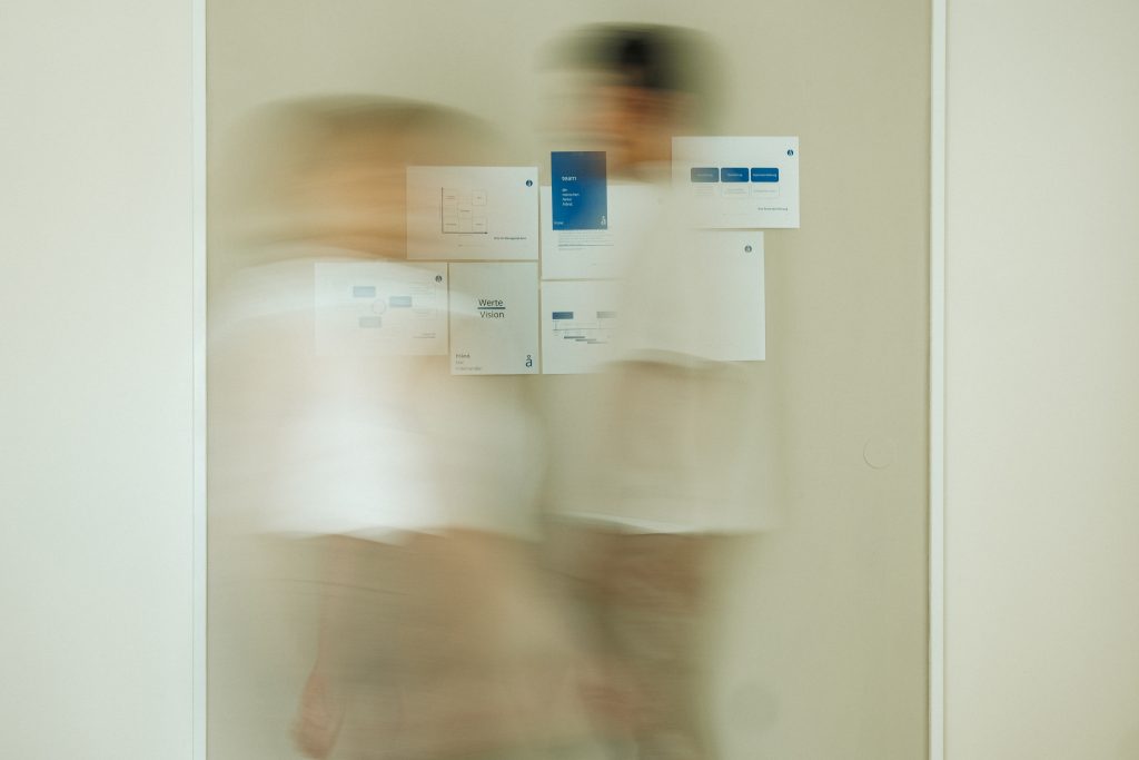 Zwei verschwommene Menschen laufen vor einer beigen Wand von rechts nach links.